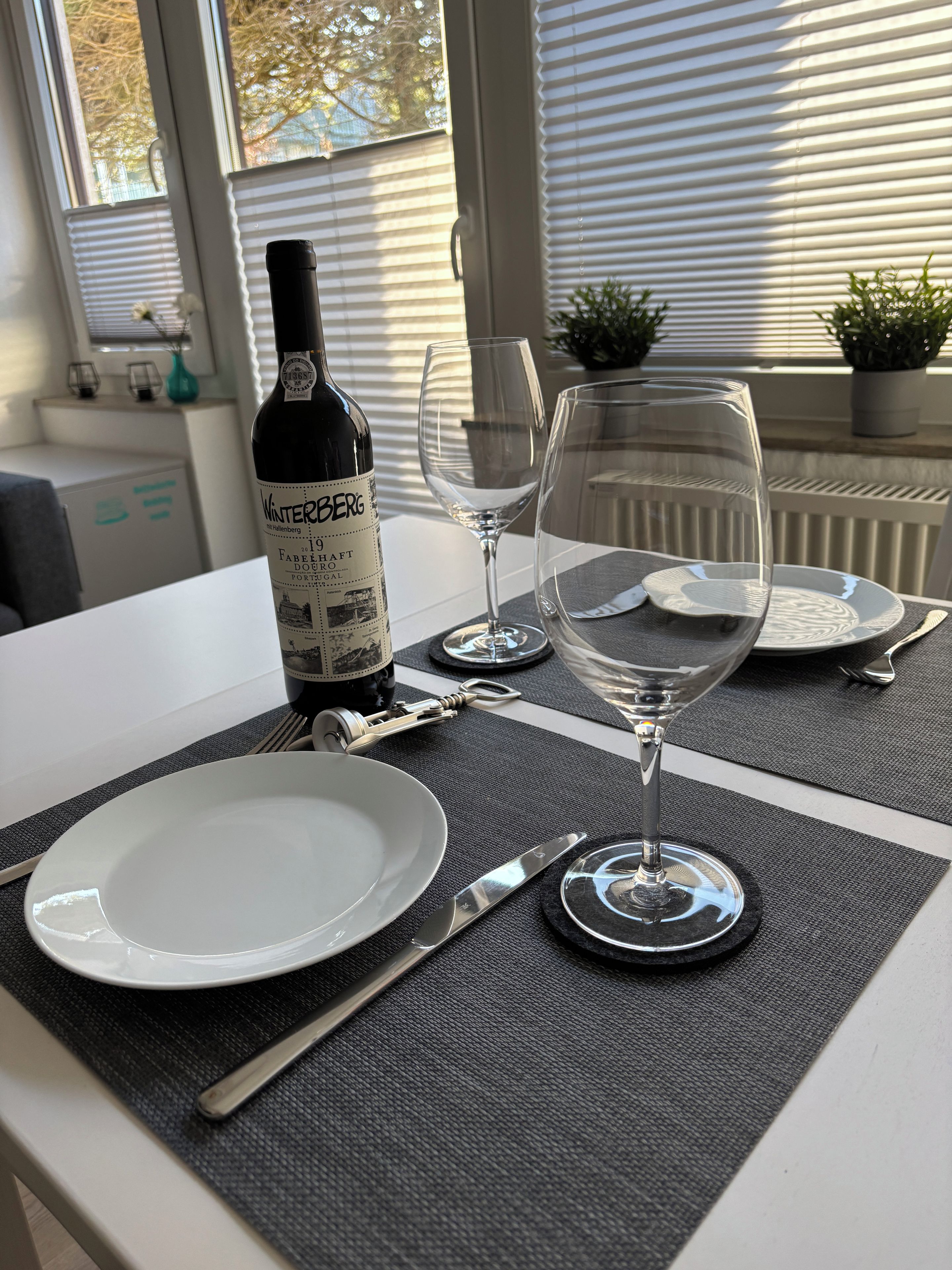 A dining table set with a bottle of wine, two wine glasses, cutlery, and plates on a textured table mat.