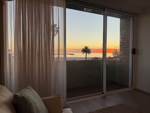 Een kamer met een schuifglazen deur die uitkomt op een balkon met uitzicht op een zonsondergang.