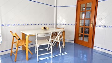 Een kamer met een betegelde muur, een houten deur, een witte tafel met stoelen en een klok aan de muur.