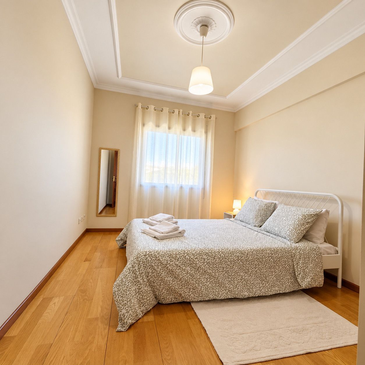 A bedroom with a bed, a bedside table, a lamp, a mirror, and a window with curtains.