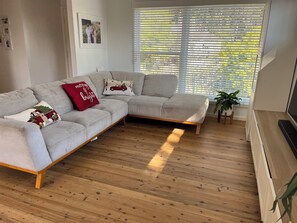 Ein Wohnzimmer mit grauem Ecksofa, Holzboden, einem TV-Schrank und einem großen Fenster mit Jalousien.