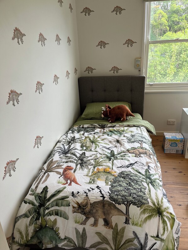 Ein Schlafzimmer mit einem Bett, das eine Bettdecke mit Dinosaurier-Motiv, ein graues Kopfteil und ein Fenster mit Blick auf Grünflächen hat.