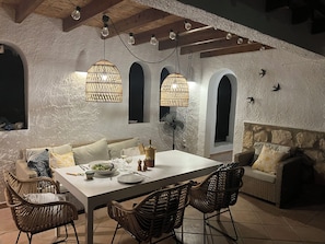 A dining area with a white table, wicker chairs, and a fan. There are two hanging light fixtures and a stone wall with arched windows. - New Listing (El Tosalet)