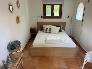 A neatly made bed with a wooden headboard, a bedside table, a small nightstand, a wicker basket, and a small stool. - New Listing (El Tosalet)