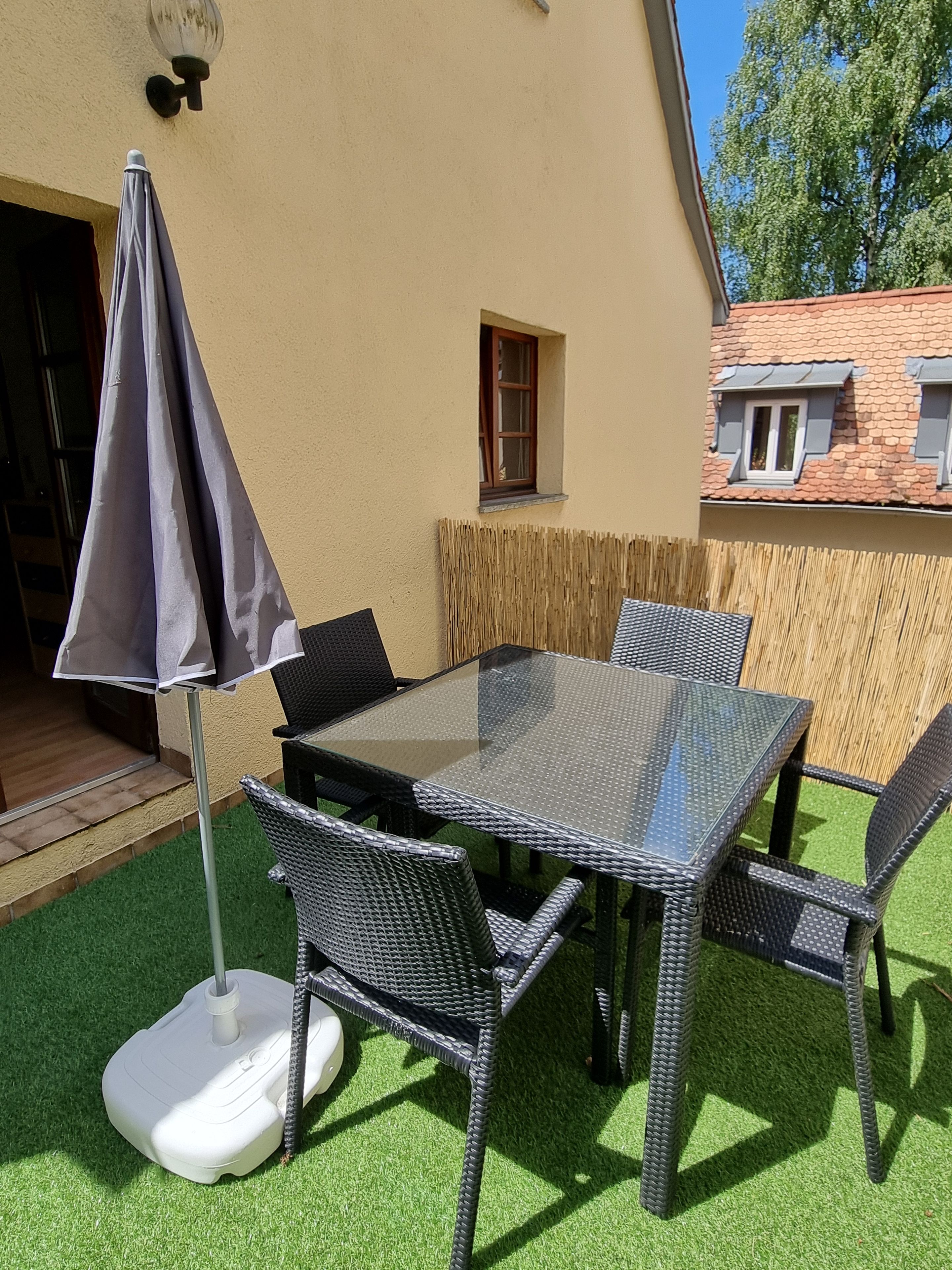 En udendørs patio med et glasbord og fletstole, omgivet af et træhegn og et lille vindue.
