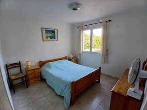 A bedroom with a single bed, a chair, a nightstand, a bedside table, a lamp, a fan, a window with curtains, and a painting on the wall. - Les Ecologies, friendly but private with space to enjoy the views (Cessenon-sur-Orb)