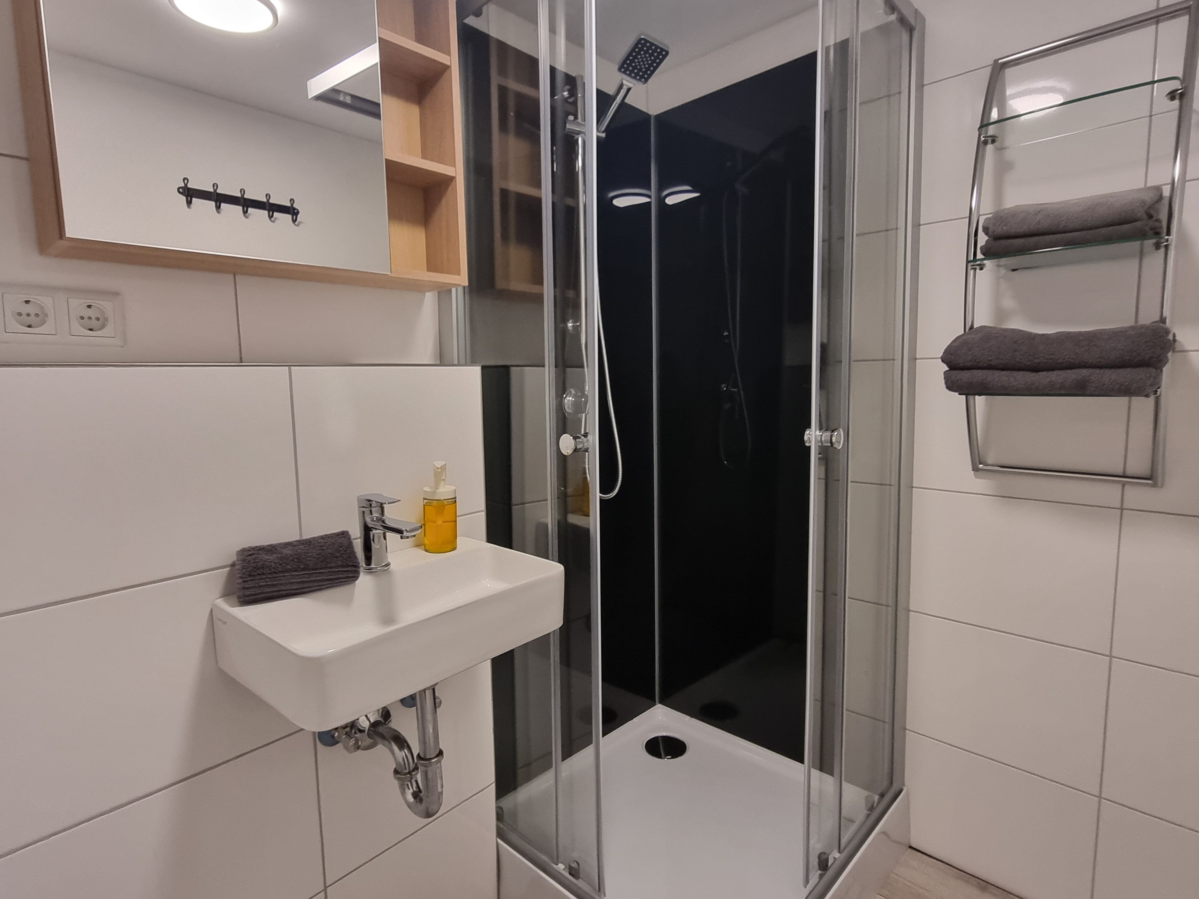 Ein modernes Badezimmer mit einer gläsernen Duschkabine, einem weißen Waschbecken inklusive Seifenspender und einem Handtuchhalter mit zwei Handtüchern.