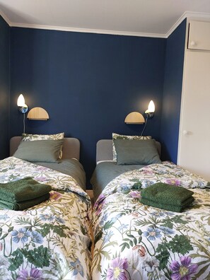 Une chambre avec deux lits, chacun recouvert d’une couvre-lit à motifs floraux et agrémenté d’un coussin vert. Des lampes murales sont fixées de chaque côté des lits.