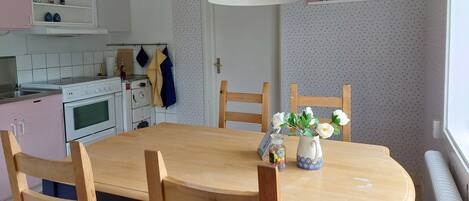 A kitchen with a dining table, chairs, a hanging light fixture, and a wall-mounted shelf with cookbooks.