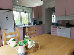 Une cuisine avec des meubles de couleur rose, un réfrigérateur blanc, une table à manger en bois ornée d’un vase de fleurs et une fenêtre avec des rideaux.