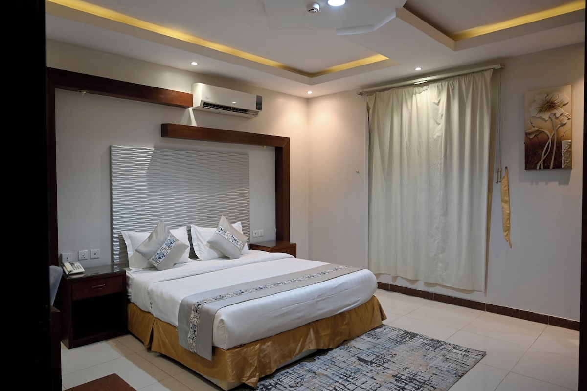 A modern bedroom with a large bed, bedside tables, a wall-mounted air conditioner, and a decorative painting.