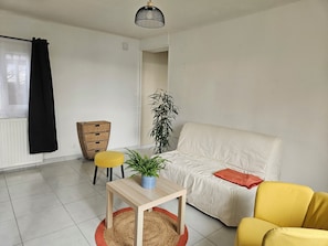 Un salon avec un canapé blanc, un fauteuil jaune, une petite table en bois ornée d’une plante et une table d’appoint en osier.