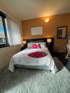 Ein Hotelzimmer mit einem Bett, einem Nachttisch, einer Lampe, einem Fenster mit Vorhängen, einem Wandspiegel und einem Teppichboden.