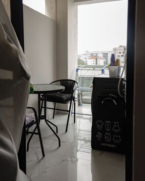 A balcony with a table and chairs, a laundry basket, and a view of buildings in the background. - Homely • Stylish • Afro-Modern • Vibrant (Nairobi)