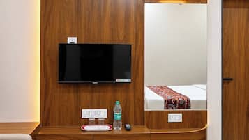 Habitación de hotel con una televisión de pantalla plana grande montada en una pared de madera, una mesita de noche con una botella de agua, un control remoto y un espejo que refleja la cama.