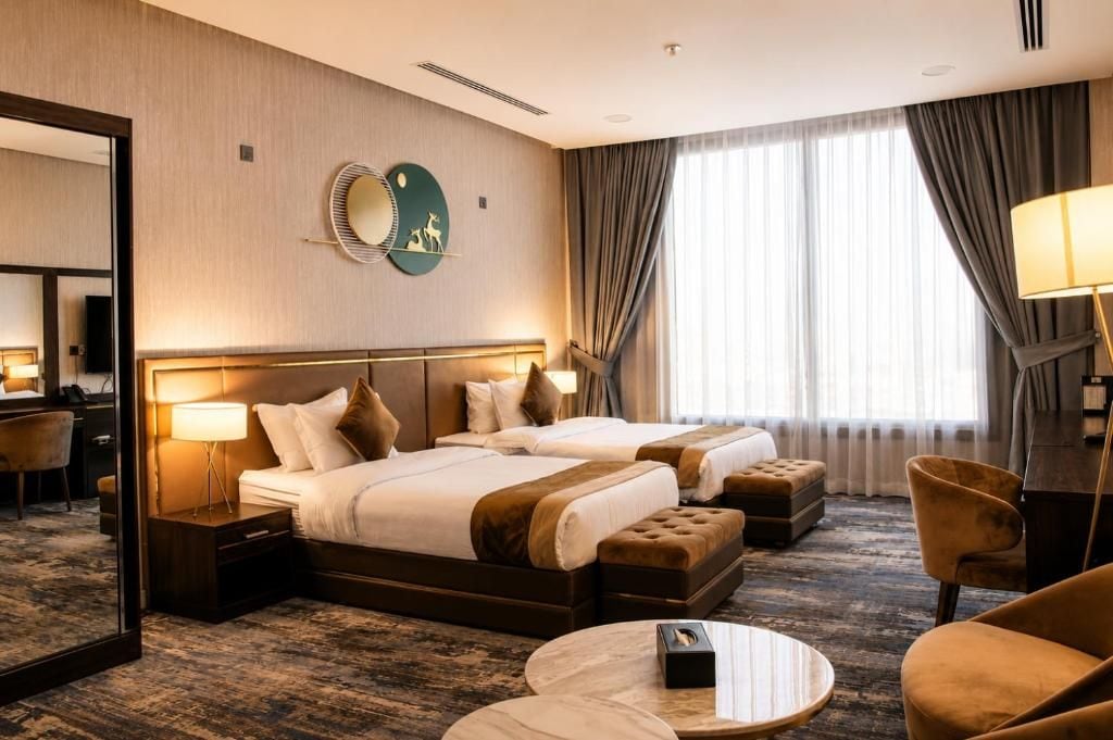 Habitación de hotel con dos camas, un espejo grande, un escritorio con lámpara y vistas al exterior a través de la ventana.