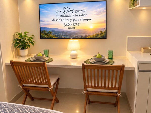 A dining area with two wooden chairs, a table with a bowl of fruit, a potted plant, and a wall-mounted TV displaying a Bible verse. - Stunning Oceanfront Condo in Santa Marta Sleeps 5 + Resort Amenities (BELLO HORIZONTE)