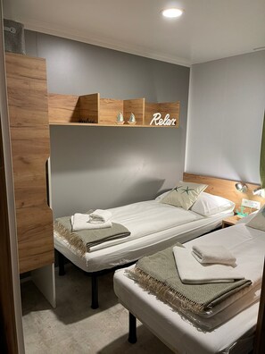 A compact hotel room with two single beds, a wooden headboard, a shelf with decorative items, and a wall-mounted light.