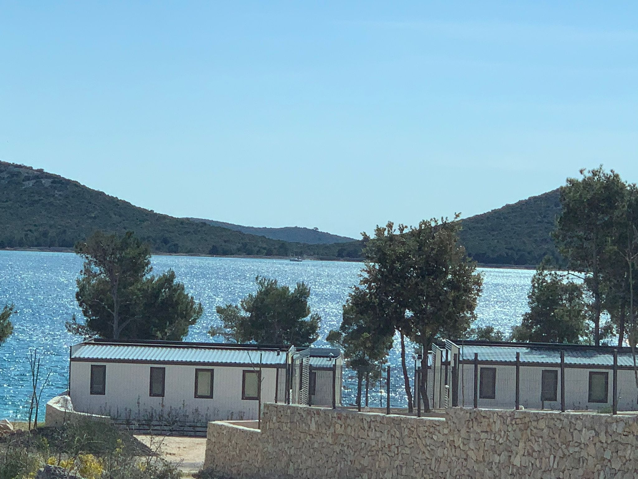 Un bâtiment situé sur la côte, avec un mur de pierre au premier plan, une étendue d’eau et des collines en arrière-plan.