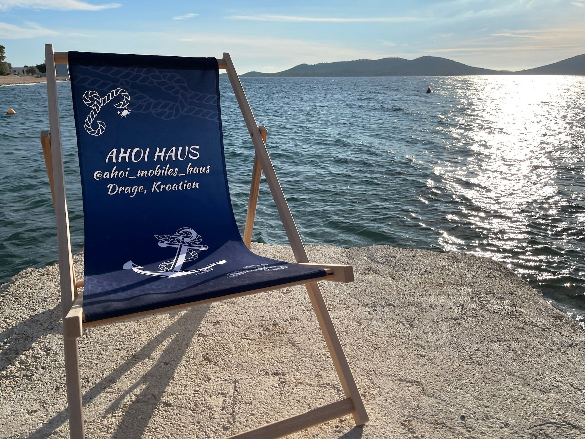 Une chaise de plage ornée d’un logo et d’un texte, placée sur une plage de galets, avec une étendue d’eau et des collines lointaines en arrière-plan.