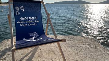 Une chaise de plage ornée d’un logo et d’un texte, placée sur une plage de galets, avec une étendue d’eau et des collines lointaines en arrière-plan.