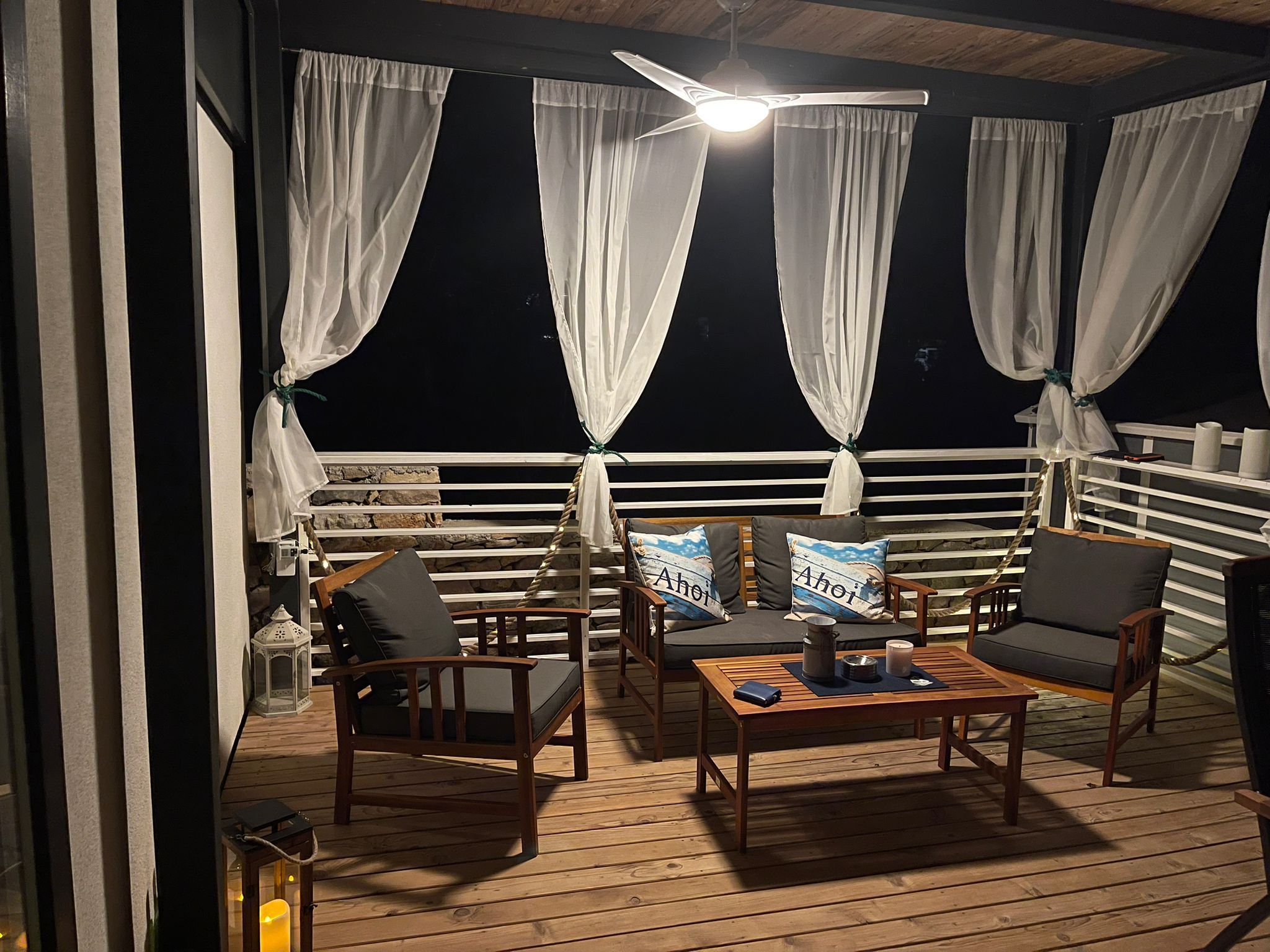 Une terrasse extérieure avec des meubles en bois, des rideaux blancs et un ventilateur de plafond.