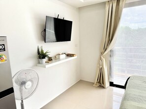 Ein modernes Wohnzimmer mit einem flachbildfernseher an der Wand, einem Standventilator, einer kleinen Pflanze und einem Fenster mit Vorhängen.
