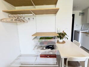 Eine kleine Küche mit einer weißen Arbeitsplatte, Holzregalen, einem Wäscheständer und einem weißen Stuhl.