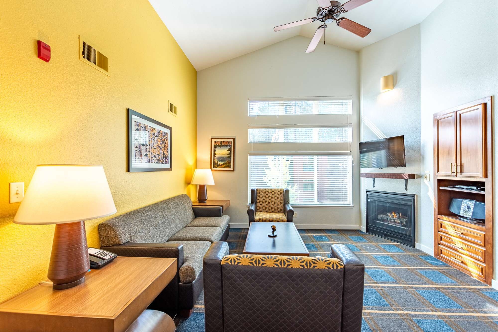 Een hotelkamer met een bank, fauteuil, tafel, lamp, plafondventilator, open haard, televisie en kunstwerken aan de wanden.