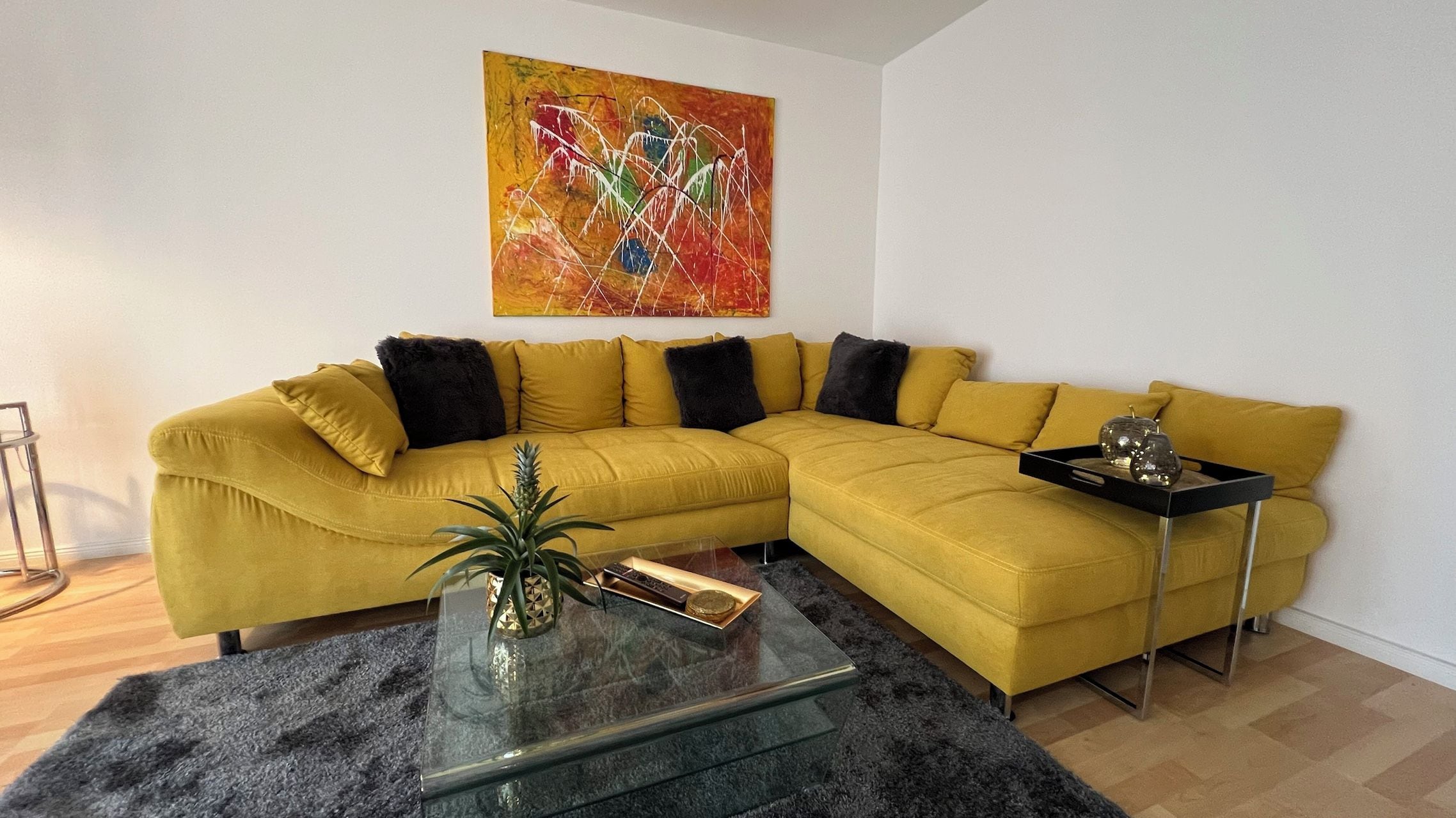 Un salon avec un canapé d’angle de couleur moutarde, une table basse en verre, une petite table d’appoint et un tableau abstrait aux couleurs vives accroché au mur.
