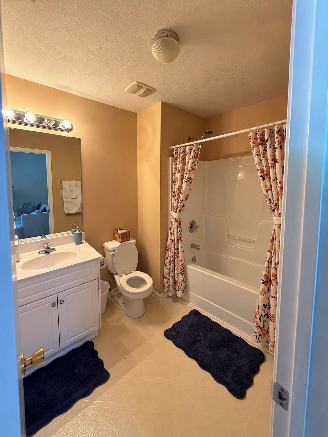 A bathroom with a sink, toilet, and bathtub, featuring a mirror with lights, a shower curtain, and a dark rug.