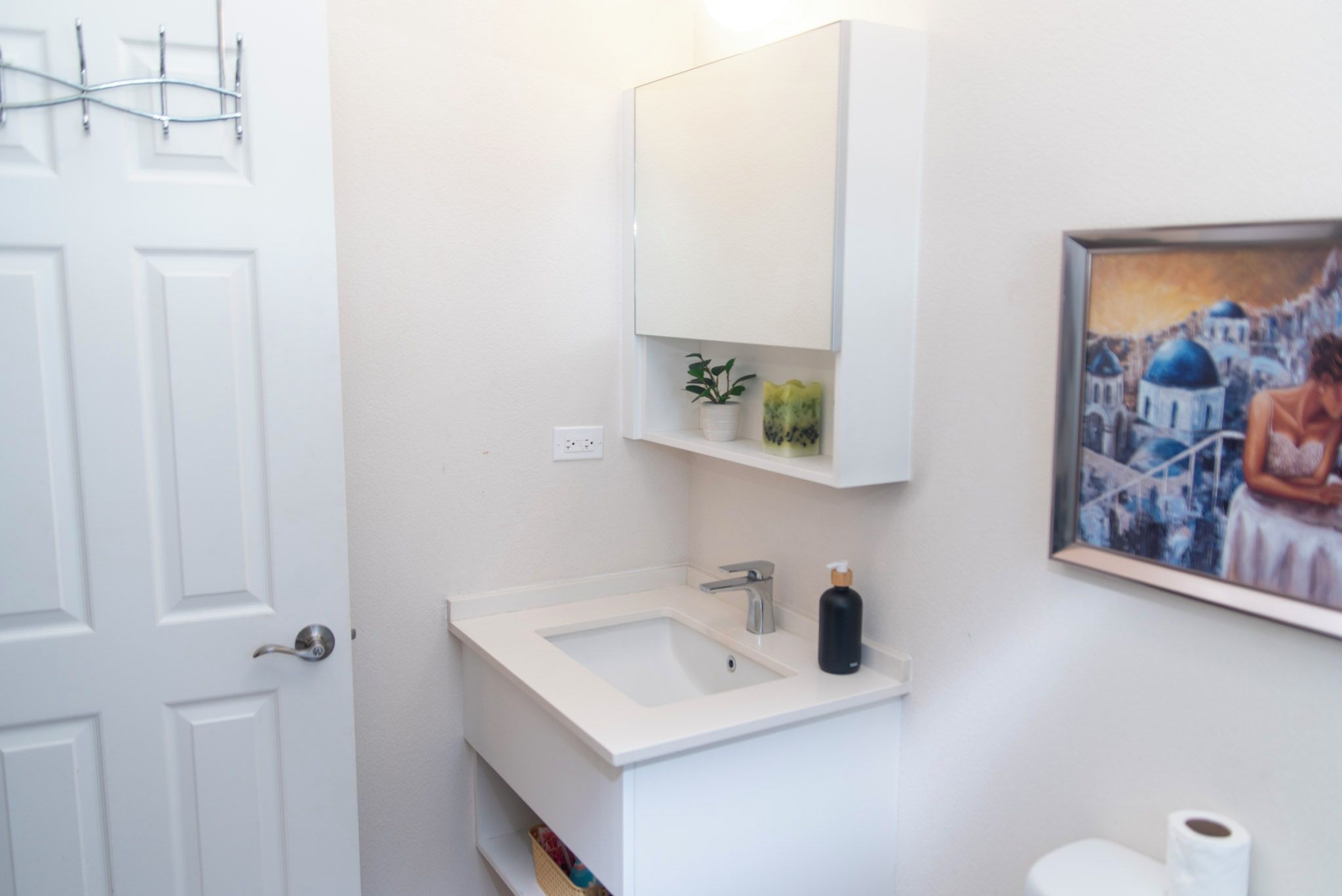 A bathroom with a white sink, a mirror, a potted plant, and a painting on the wall.