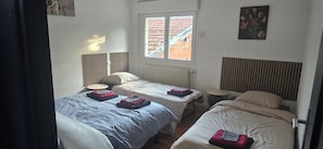 A room with two single beds, a small round table, a radiator, and a window with a view of buildings. - New Listing (Ville-sur-Saulx)