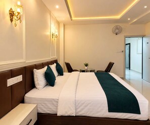 A hotel room with a bed, bedside table, lamp, wall-mounted light, and a small table with a plant. - Suite Room Hotel Ashok Vatika (Nagpur)