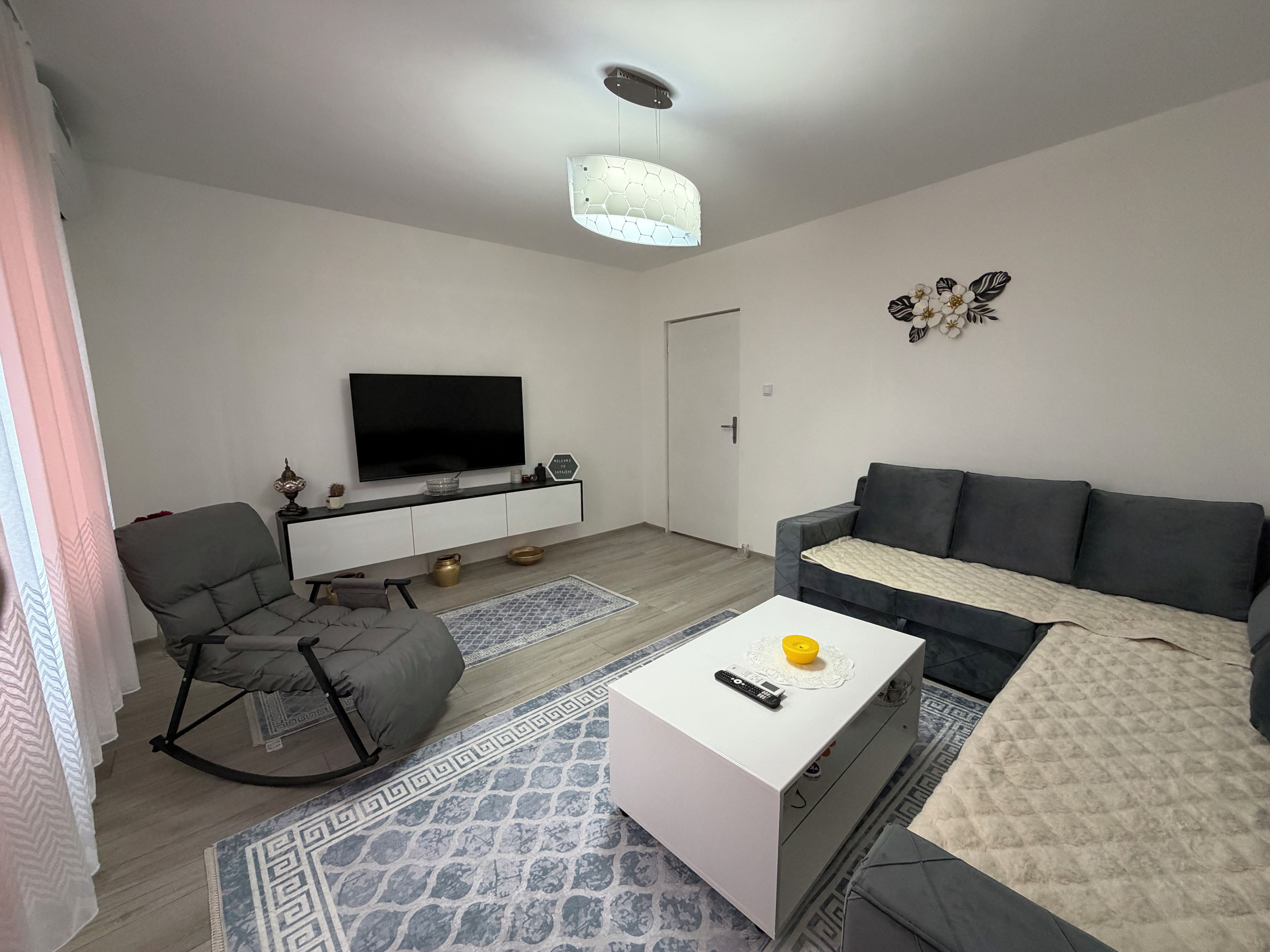 A modern living room with a flat-screen TV, grey sofa, white coffee table, and patterned rugs.