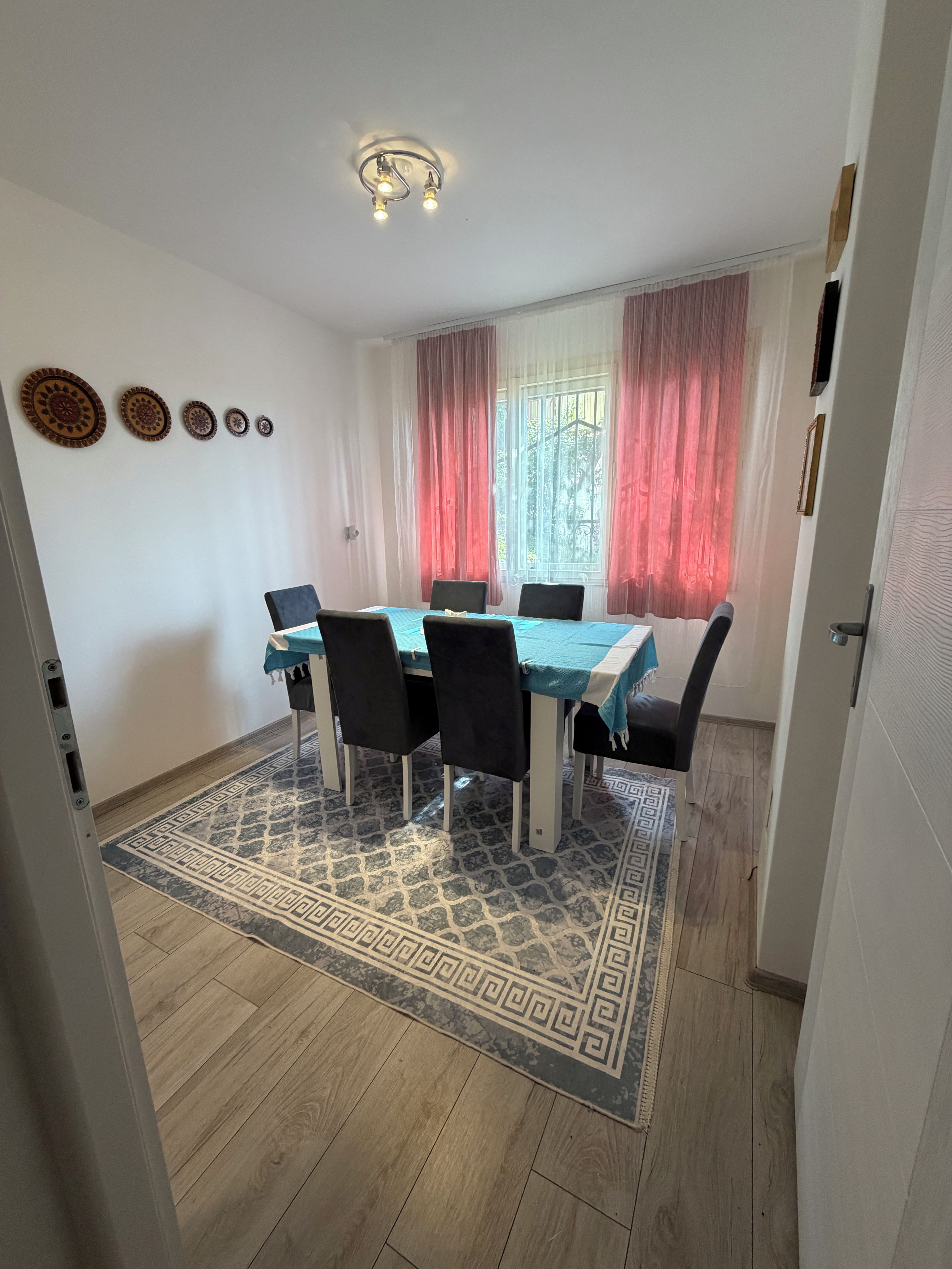 A dining area with a table, chairs, a patterned rug, and a window with curtains.