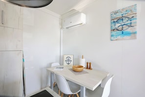 A small, white-walled room with a white ceiling, a white table, and a white chair. There is a white air conditioning unit mounted on the wall, a wooden bowl on the table, and a wooden chest with a label. - Ahoi House 10 – Seaside mobile home in Drage, sleeps 4 (Drage)