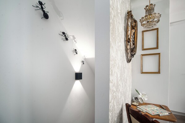 Une pièce avec un mur blanc, une chaise en bois, une table sur laquelle est posé un jeu de société, ainsi que des objets décoratifs, notamment des sculptures de fourmis et un lustre.