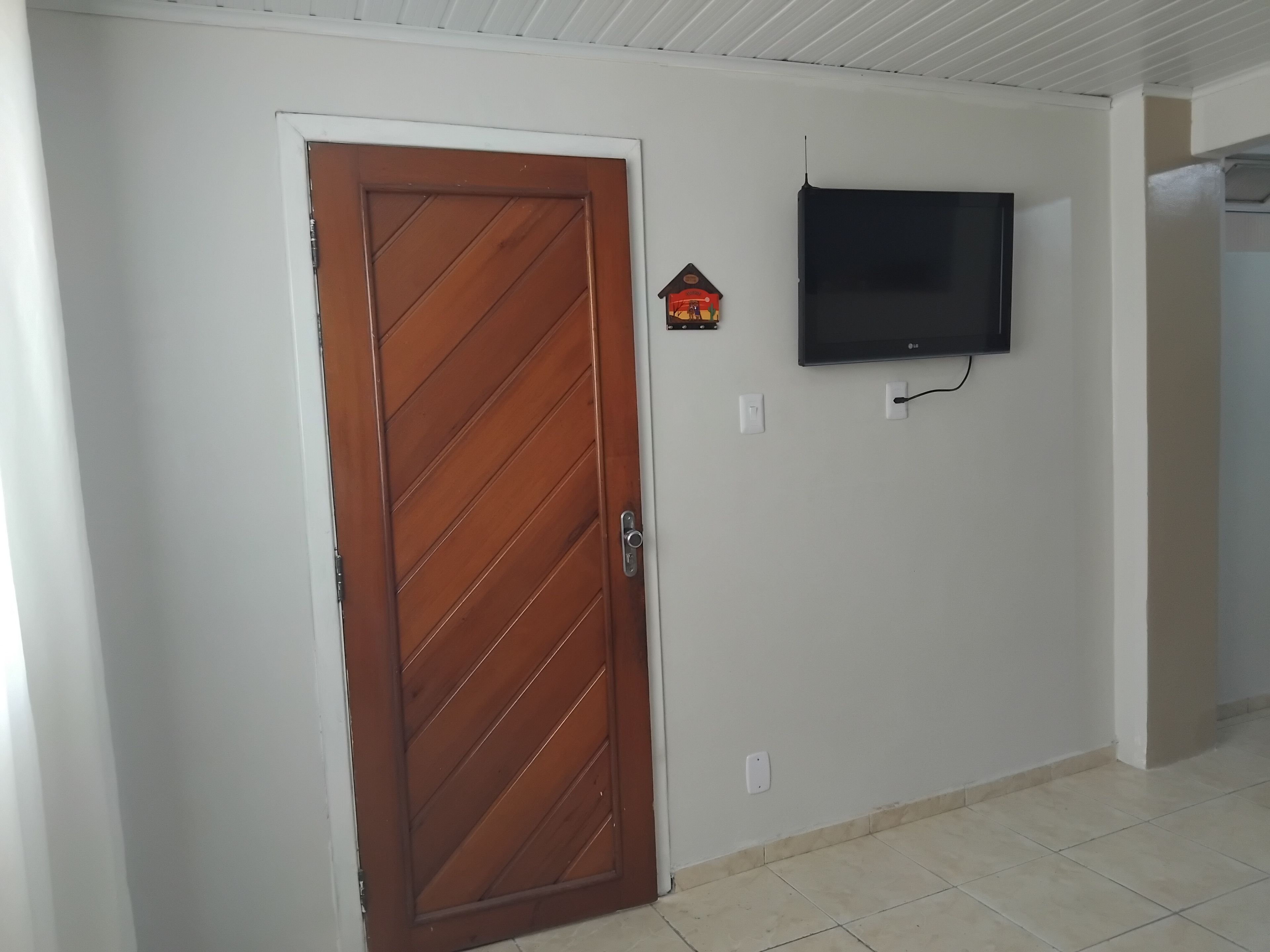 Una habitació amb una porta de fusta, televisió penjada a la paret i un terra amb rajoles.