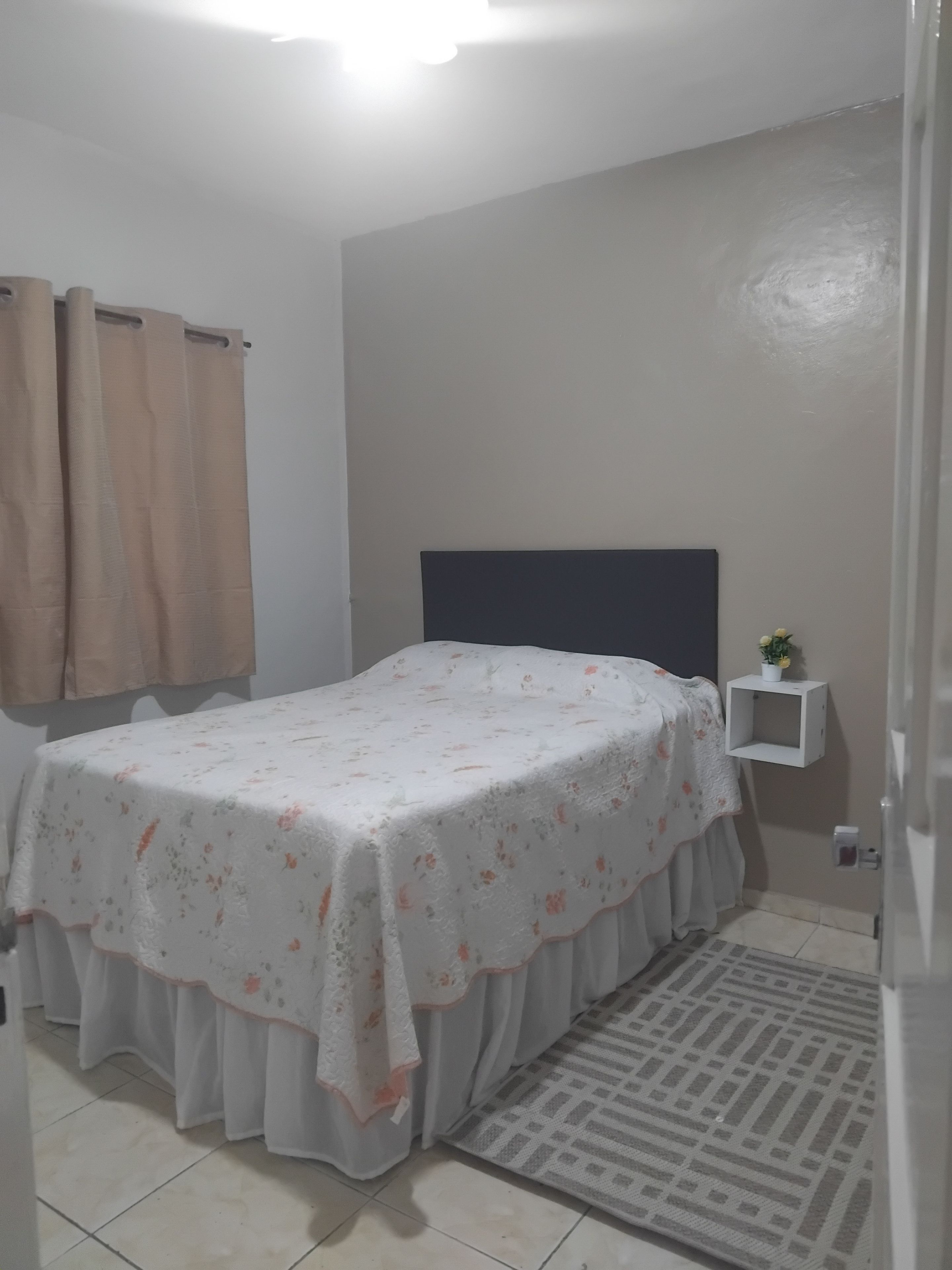 Un dormitorio con cama, cortinas, una mesita de noche con una planta y una pequeña estantería.