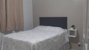 Un dormitorio con cama, cortinas, una mesita de noche con una planta y una pequeña estantería.