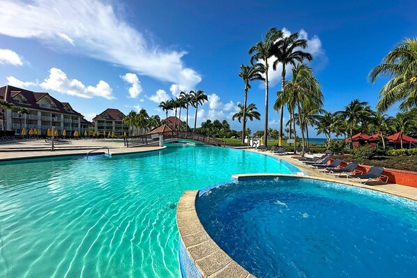 A large swimming pool with lounge chairs and palm trees, surrounded by hotel buildings. - New Listing (Sainte-Luce)