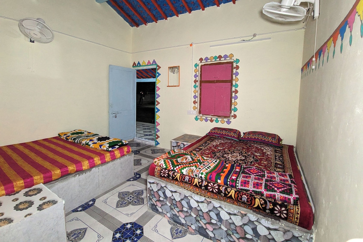 A room with a bed, a small table, a fan, and a window with a decorative frame.