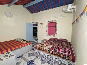 A room with a bed, a small table, a fan, and a window with a decorative frame. - White Rann Homestay (Dhordo)