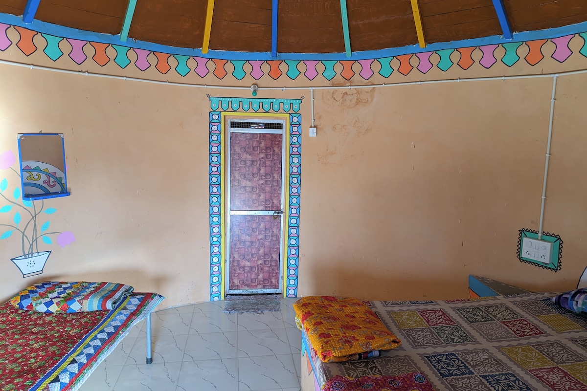 A room with a tiled floor, a bed with a colorful quilt, a small table with a plant, and a door with a decorative frame.