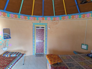 A room with a tiled floor, a bed with a colorful quilt, a small table with a plant, and a door with a decorative frame. - White Rann Homestay (Dhordo)