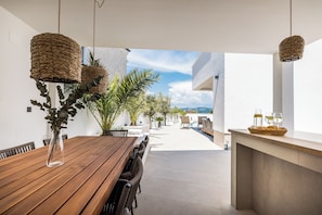 Un espacio moderno para comer al aire libre con una mesa y sillas de madera, lámparas de mimbre colgantes y plantas en macetas.