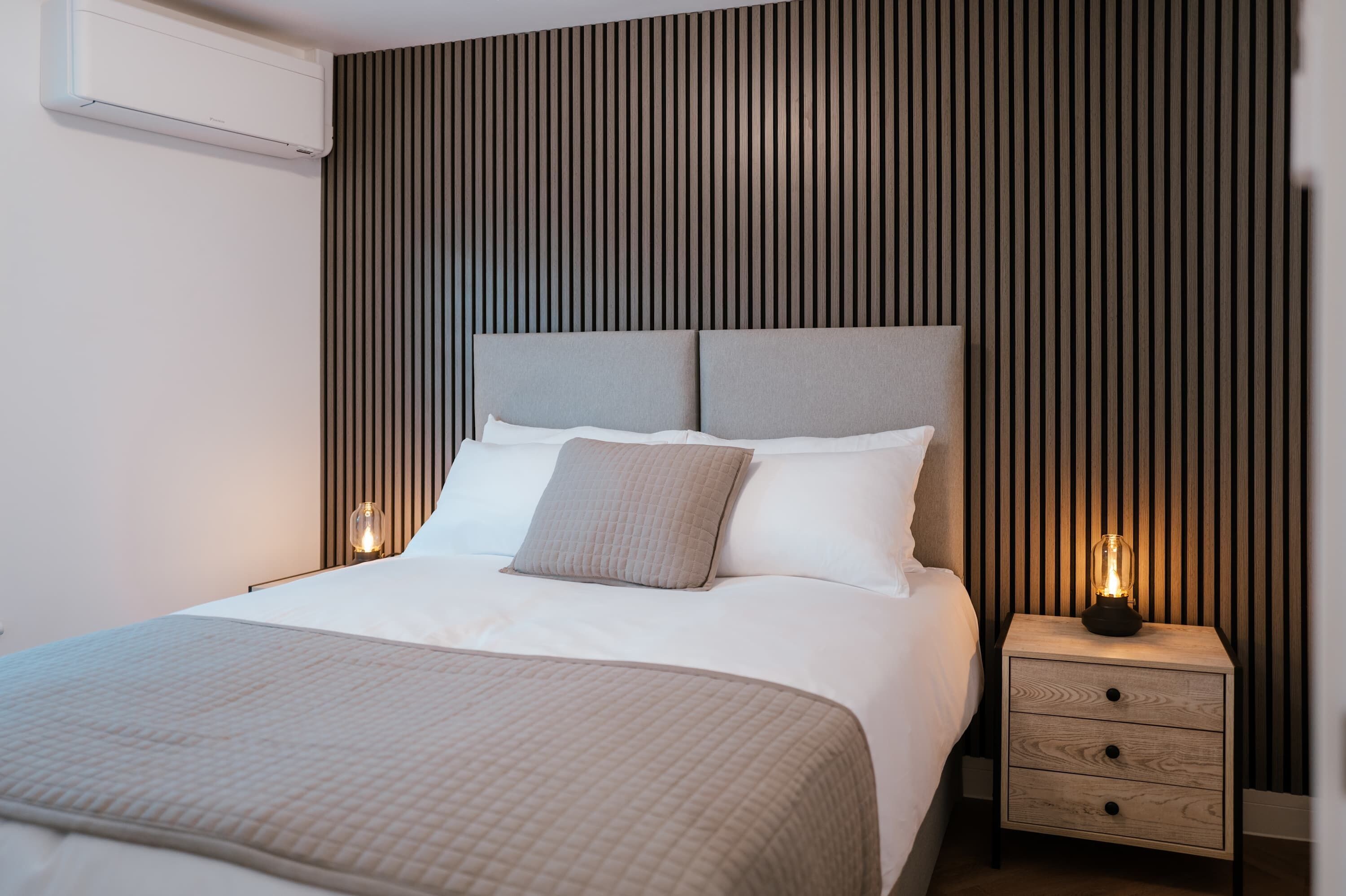 A modern hotel room with a large bed, a bedside table with a lamp, and a wall-mounted air conditioner.