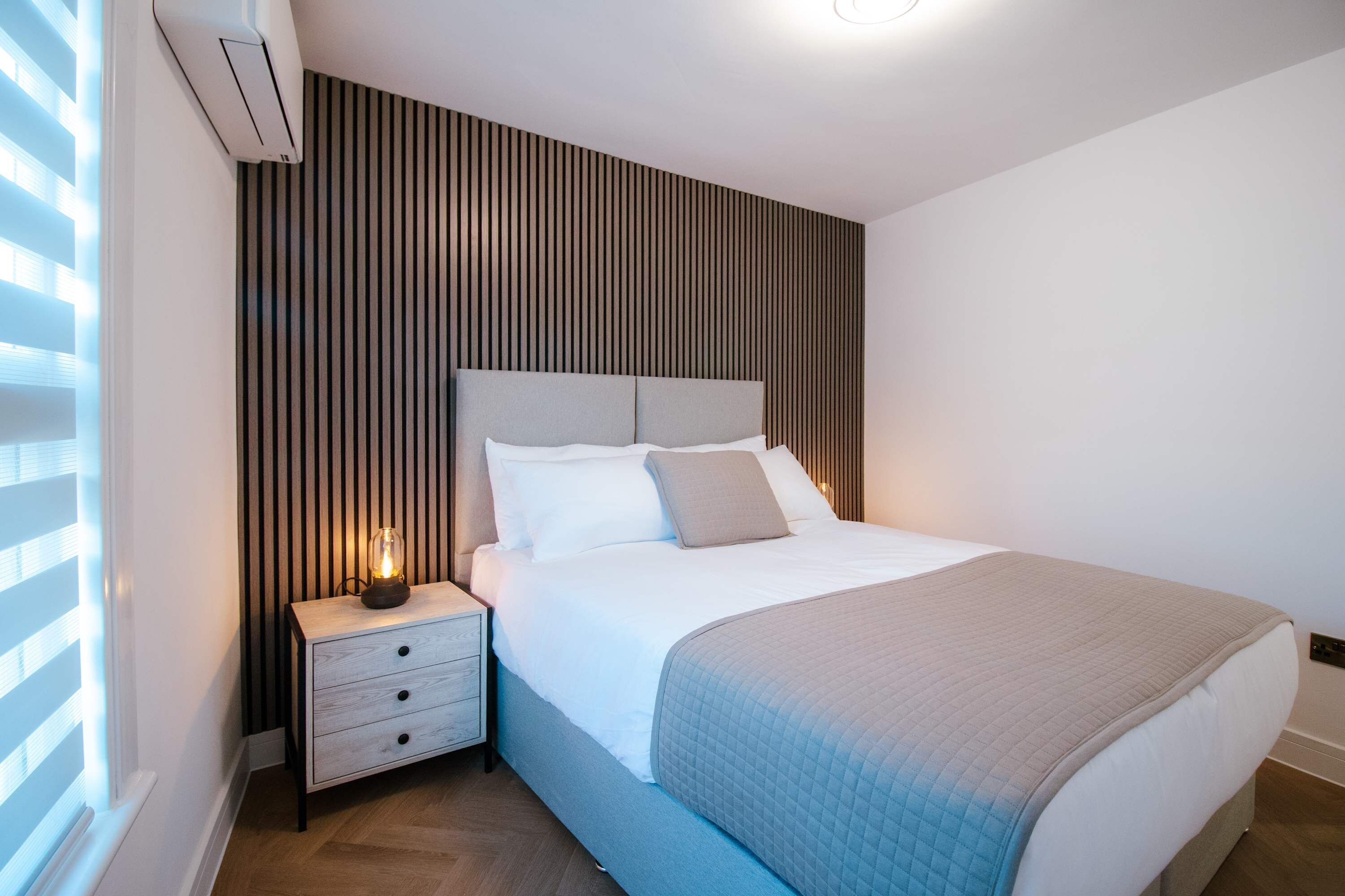 A modern bedroom with a bed, nightstand, and a wall-mounted air conditioning unit.