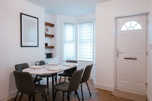 A dining area with a white table set for four, surrounded by chairs. There is a framed quote on the wall, a window with blinds, and a white door with a handle. - Perfect Booking 2-bed Near Folkestone Harbour (Folkestone)